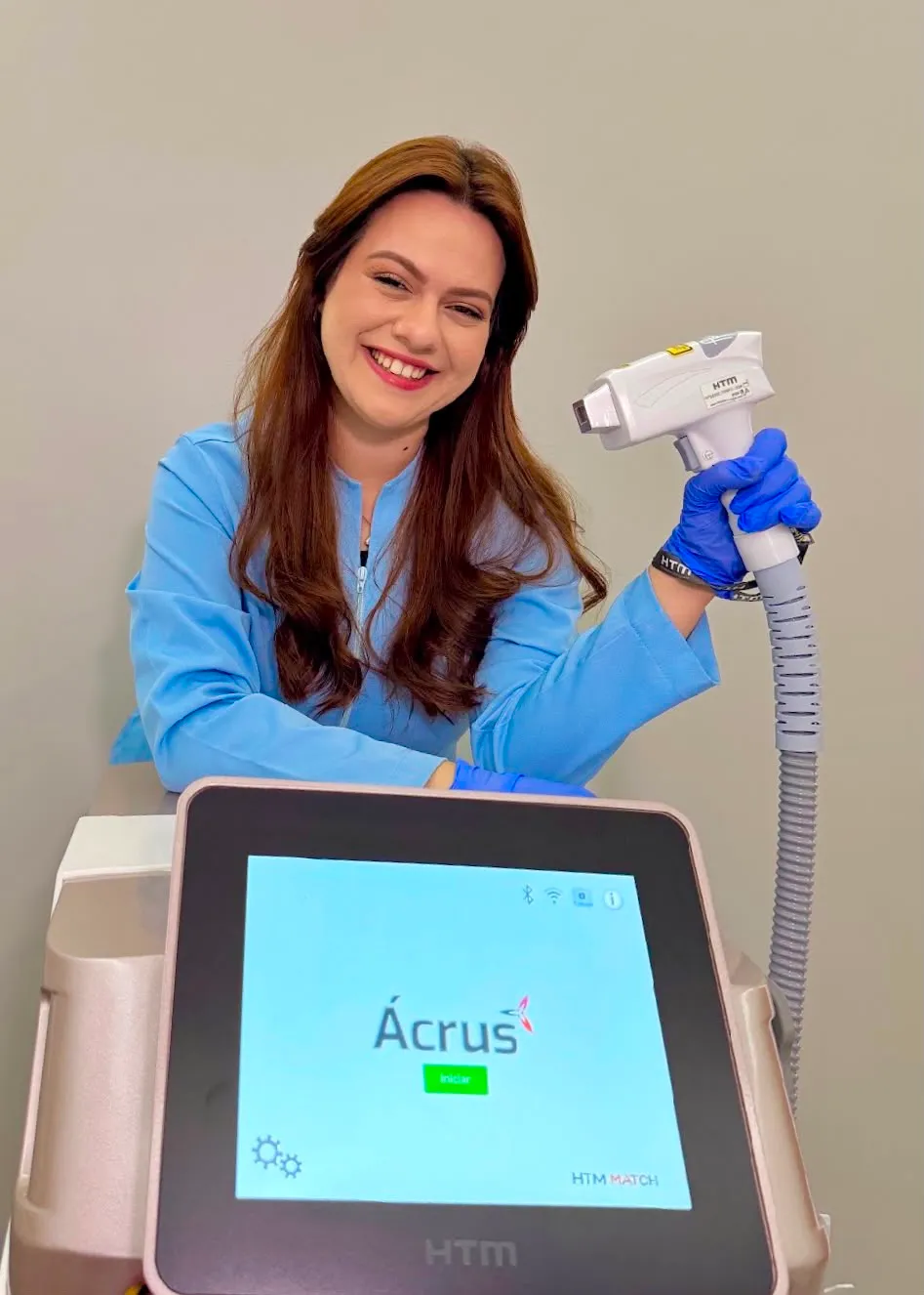 Fabiane, responsável pela depilação a laser, de jaleco azul e luva, com a ponteira do laser em mãos ao lado do aparelho